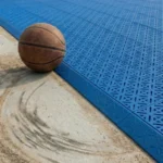 砂埃で滑った跡が残るコンクリートの地面と、雪の結晶パターンが施された青いNEXCOURTスポーツタイル床の比較写真。境目にバスケットボールが置かれ、タイルの端にはスロープと専用のコーナー連結パーツが設置されている。