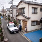 日本の住宅密集地の狭い道にパトカーが停まり、警察官が玄関先で住人と話している。右手前の敷地には車1台分ほどの青いスポーツタイルのコートとバスケットゴールが1基ある