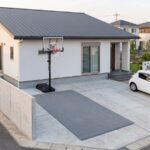 ガルバリウム鋼板の屋根と白い塗り壁の新しい日本の住宅。2台分のコンクリート駐車場で、右の1台分に白い普通乗用車が停まり、左の1台分だけに濃いグレーのインターロッキング式スポーツタイルが細長く敷かれている。タイルの表面は単色の無地で、パネルの継ぎ目のグリッドだけが見えている。タイルの範囲は隣の車と同じ幅・同じ長さで、幅2.5m×奥行6.0mの1台分。奥の壁際にタンク式バスケットゴールが立ち、黒いベースはタイルの外のコンクリートに置かれている
