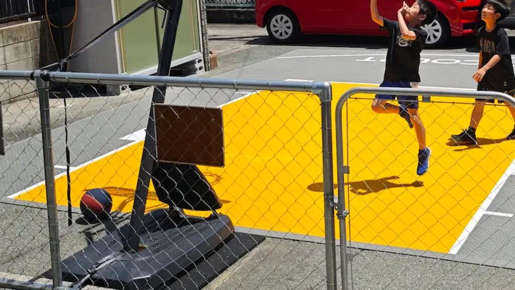 タンク式バスケットゴールを置いた駐車場のコート。ゴールのベースはタイルの外側の舗装面にあり、その前に黄色とグレーのタイルが敷かれていて、子供がシュートを打っている