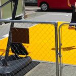 タンク式バスケットゴールを置いた駐車場のコート。ゴールのベースはタイルの外側の舗装面にあり、その前に黄色とグレーのタイルが敷かれていて、子供がシュートを打っている