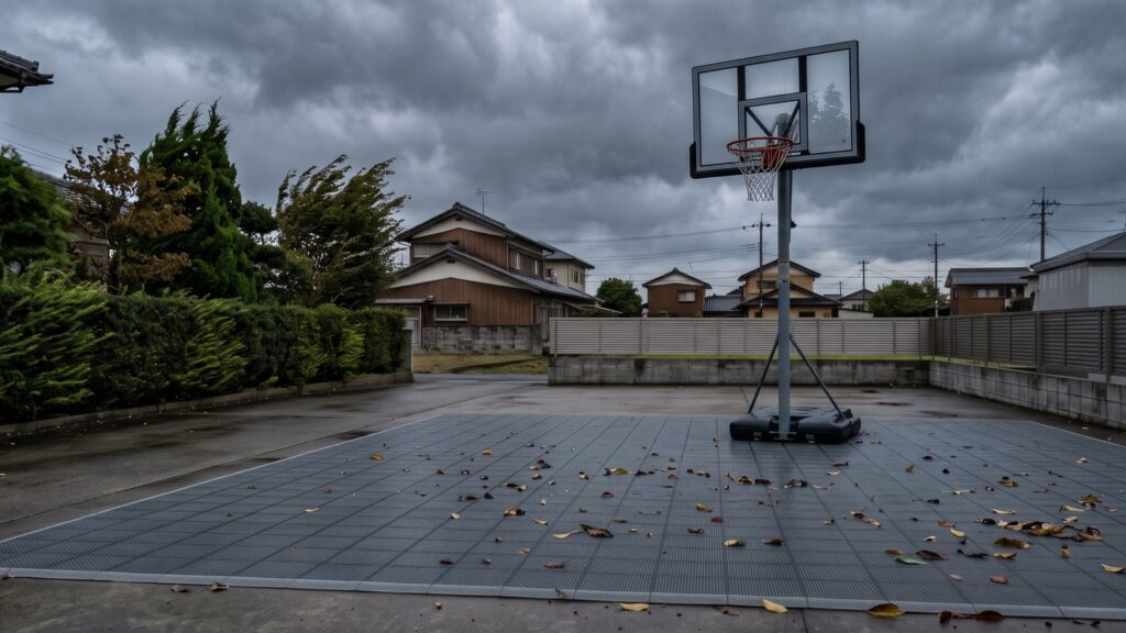 台風が接近する夕方の日本の住宅の駐車場。インターロッキング式スポーツタイルのコートに落ち葉が吹き寄せられ、奥にタンク式バスケットゴールが立っている。黒い水タンクのベースから垂直にマストが伸び、斜めのブレースで三角に支えられ、上端で前へ持ち出されたバックボードにオレンジのリングとネットが付いている。空は分厚い鉛色の雨雲で、庭木が強風で同じ方向にたわんでいる