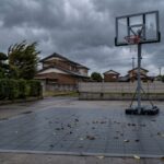 台風が接近する夕方の日本の住宅の駐車場。インターロッキング式スポーツタイルのコートに落ち葉が吹き寄せられ、奥にタンク式バスケットゴールが立っている。黒い水タンクのベースから垂直にマストが伸び、斜めのブレースで三角に支えられ、上端で前へ持ち出されたバックボードにオレンジのリングとネットが付いている。空は分厚い鉛色の雨雲で、庭木が強風で同じ方向にたわんでいる