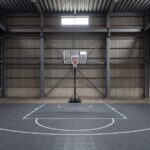 日本の鉄骨造倉庫の内部。正面の壁際にタンク式バスケットゴールが立ち、その壁に沿って左右いっぱいに濃いグレーのインターロッキング式スポーツタイルが柱スパンいっぱいに敷き詰められている。白いラインで制限区域とフリースロー半円、アークが引かれ。高い天井には鉄骨の梁が露出している